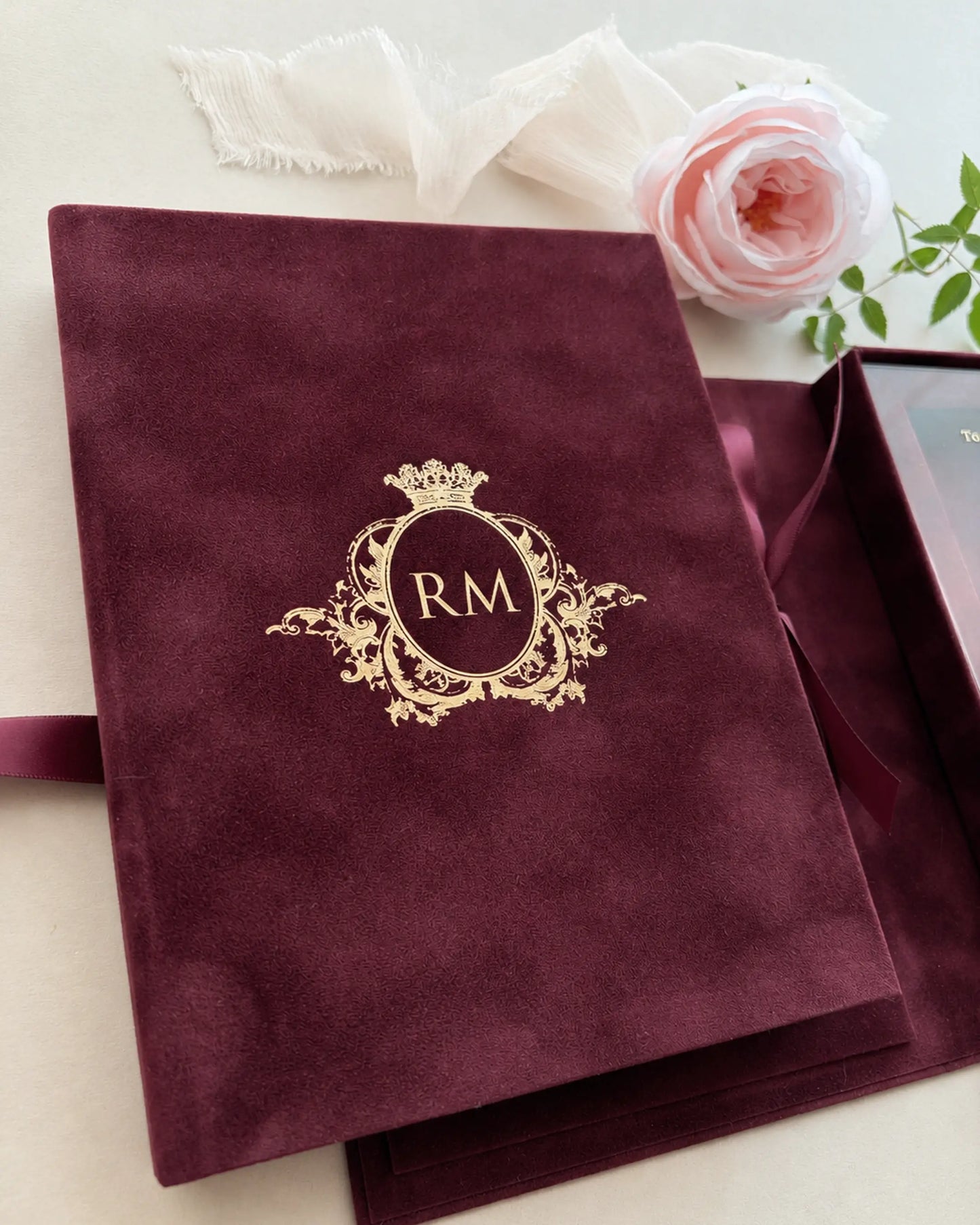 Macro shot of a luxurious burgundy velvet wedding invitation book cover featuring an intricate regal crest and custom initials pressed in reflective gold foil — Polina Perri