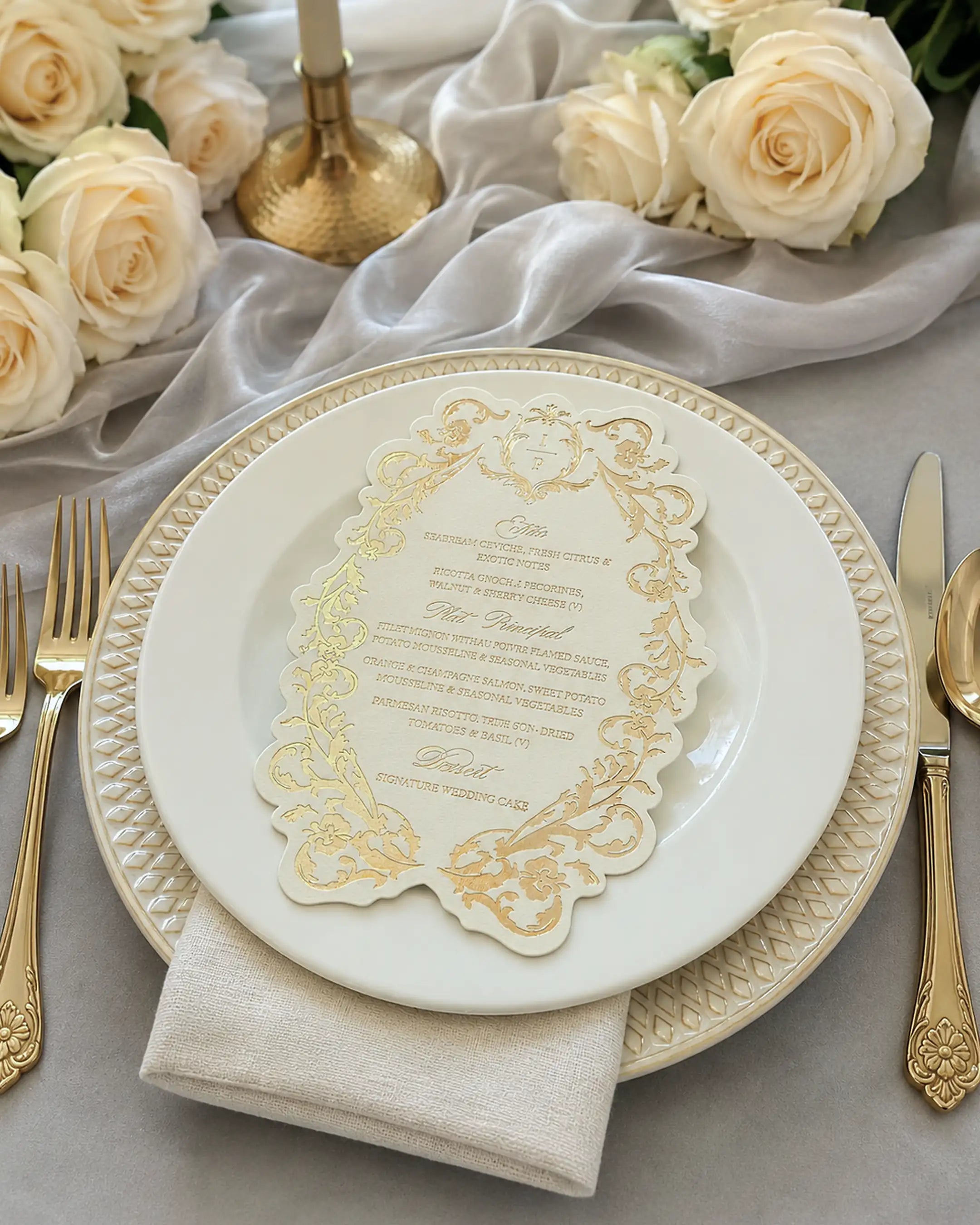 An elegant wedding reception table setting showcasing a die-cut ornate menu card with intricate gold foil patterns, placed on a white porcelain charger plate surrounded by white roses and luxury gold cutlery — Polina Perri