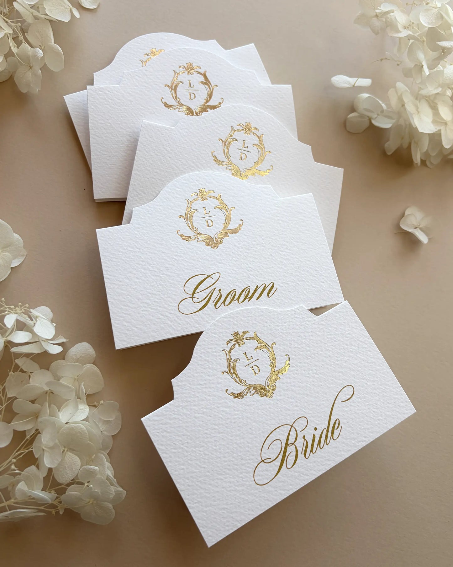 Elegant Arch Place Cards with gold foil monogram scattered on beige background with white hydrangeas — Polina Perri