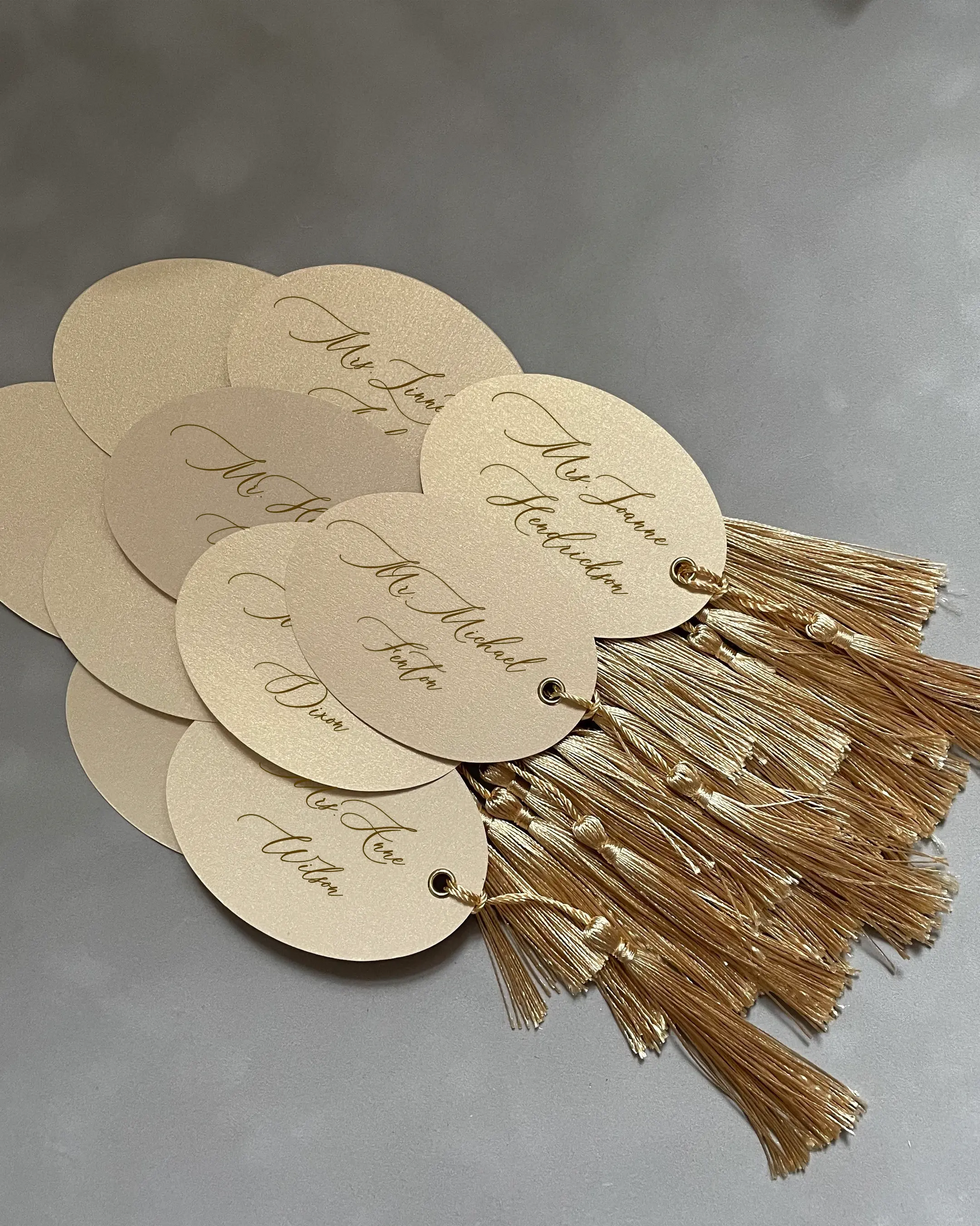 Cream Oval Place Cards with gold tassels and calligraphy script names fanned out together — Polina Perri