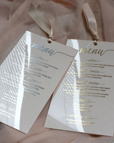 Two clear acrylic wedding menu cards side-by-side demonstrating the choice between shiny gold foil and metallic silver foil printing with matching silk ribbons — Polina Perri