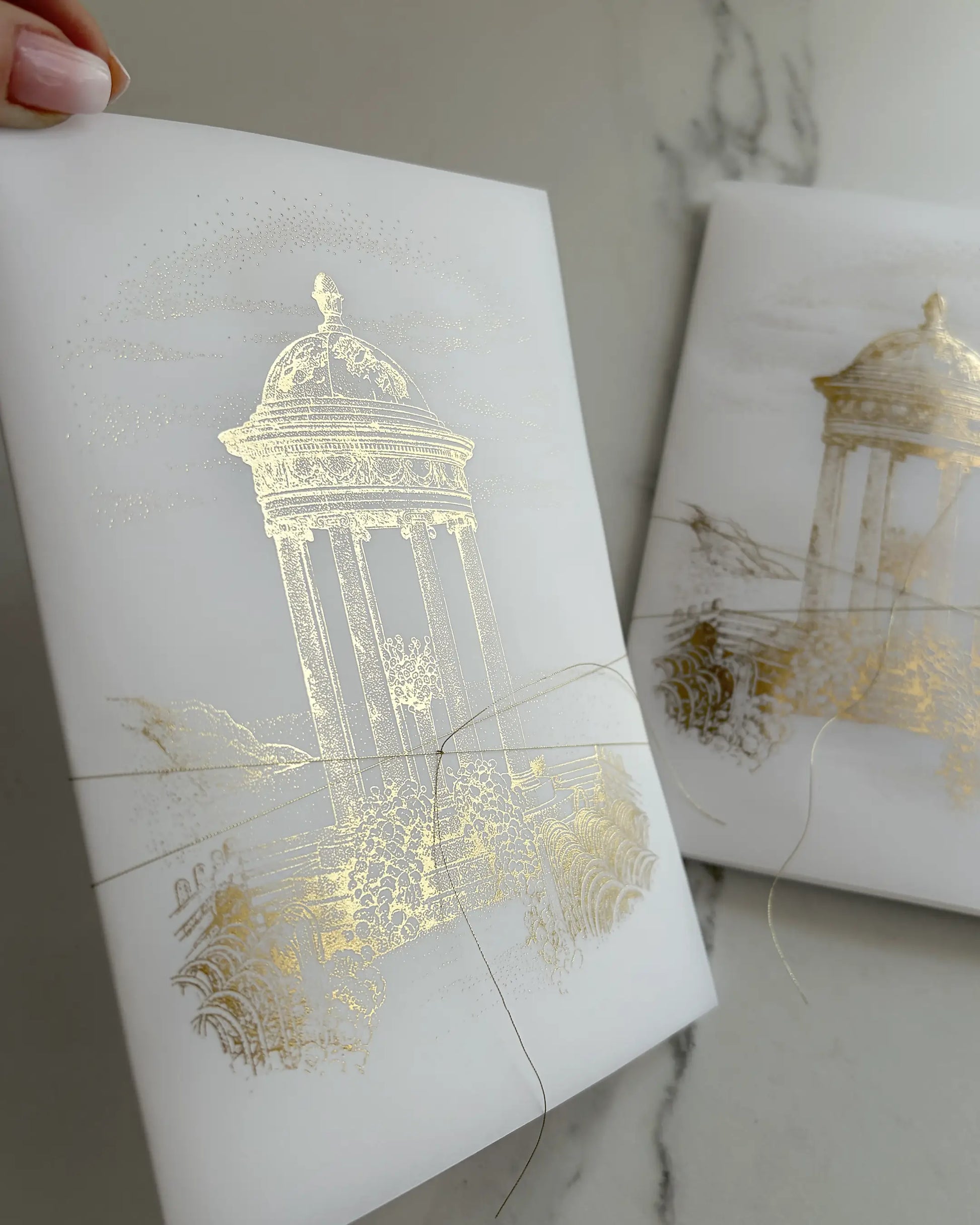 Belvedere luxury destination wedding invitations close up of semi transparent tri fold vellum wrap with gold foil classical pavilion design and delicate gold string Polina Perri