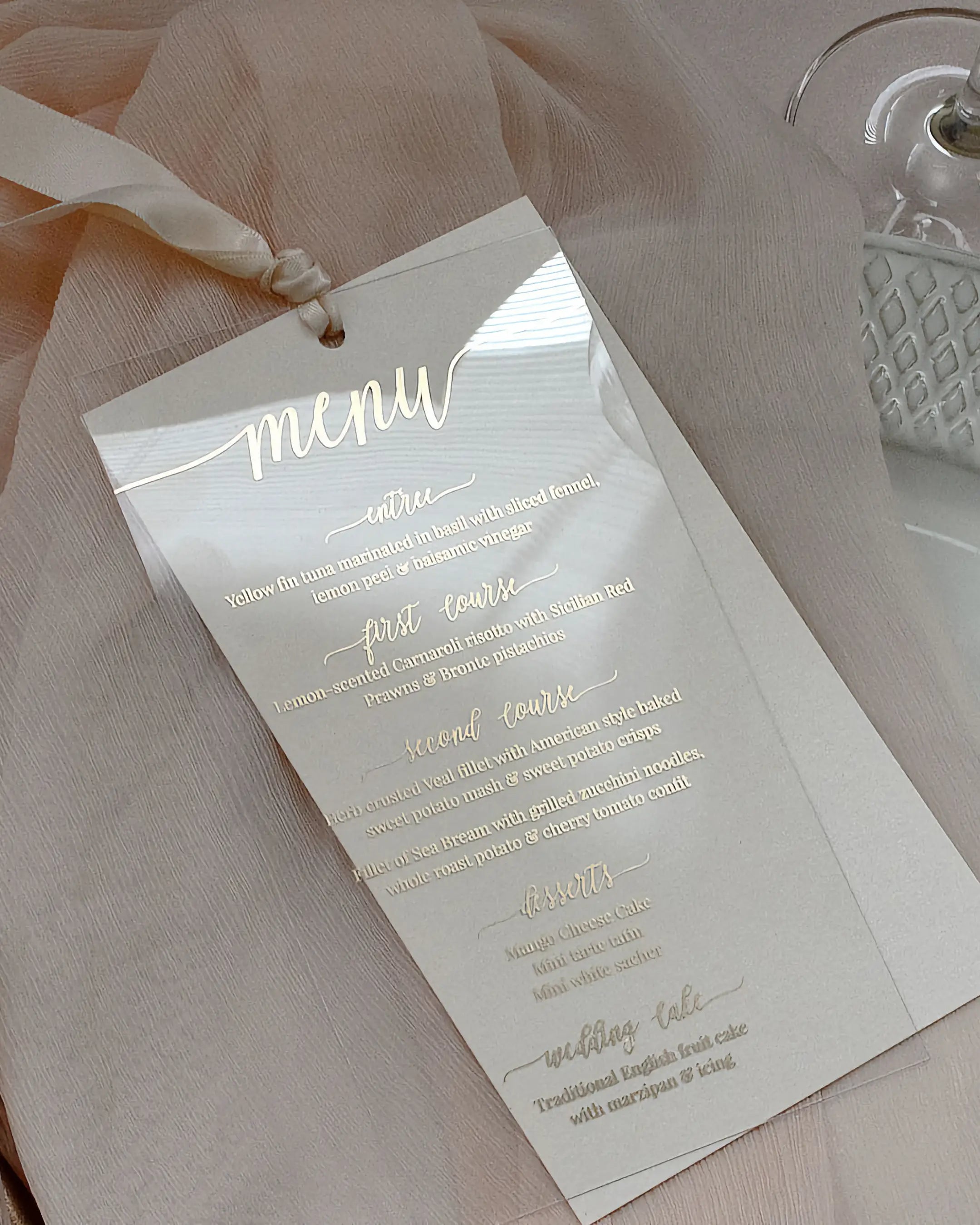 Modern clear acrylic wedding menu card featuring elegant gold foil script typography and a luxury silk ribbon on a soft pink fabric background — Polina Perri