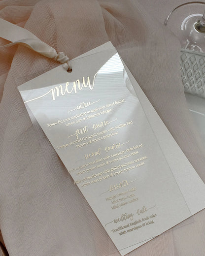 Modern clear acrylic wedding menu card featuring elegant gold foil script typography and a luxury silk ribbon on a soft pink fabric background — Polina Perri