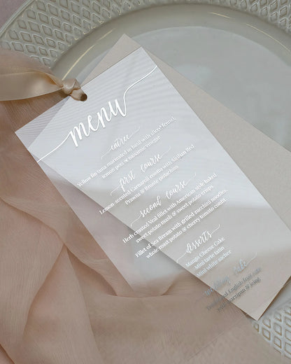 Contemporary wedding table setting featuring a clear acrylic menu card with striking silver foil script positioned on a white textured charger plate — Polina Perri