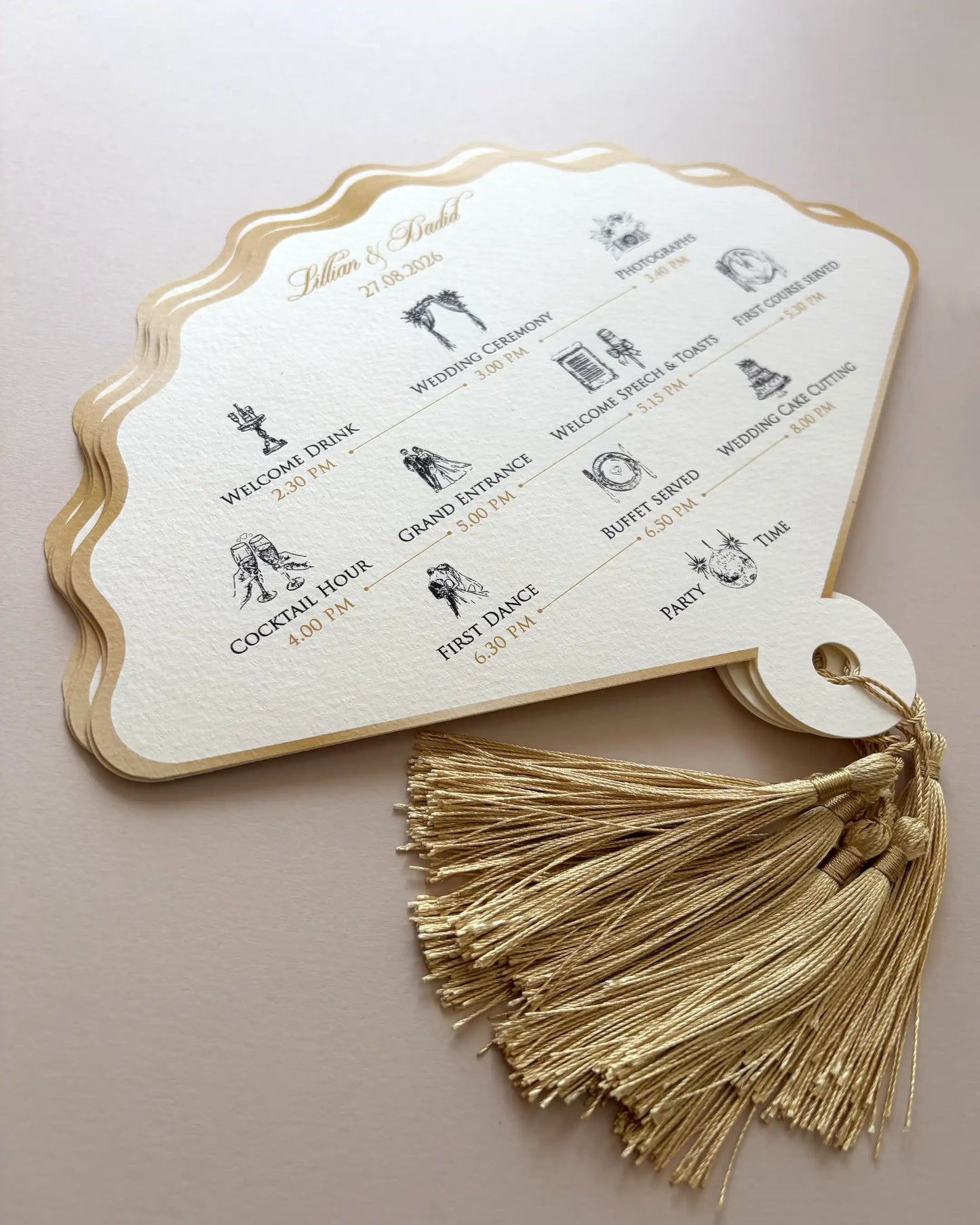 A focused close-up photo of a luxury fan wedding order of the day printed on layered cream paper with detailed icons, gold foil edges, and an intricate gold tassel, against a clear neutral background — Polina Perri