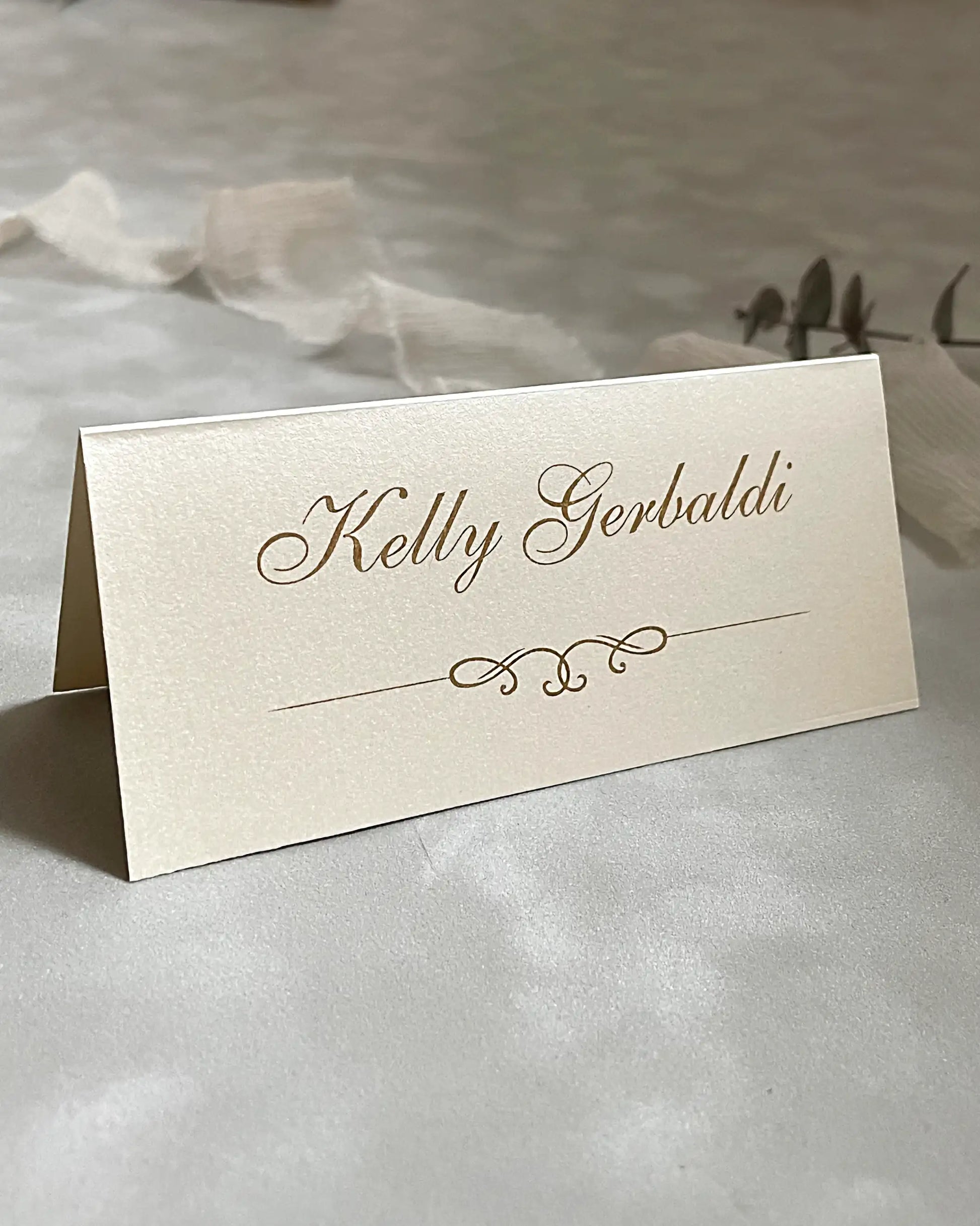 Classic cream shimmer wedding tent place card featuring elegant cursive typography and a delicate hot foil pressed gold swirl motif — Polina Perri