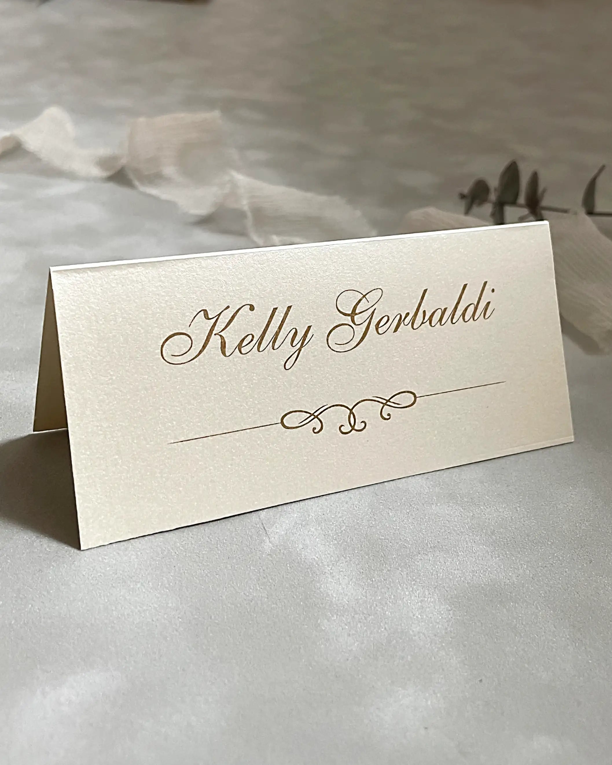 Classic cream shimmer wedding tent place card featuring elegant cursive typography and a delicate hot foil pressed gold swirl motif — Polina Perri