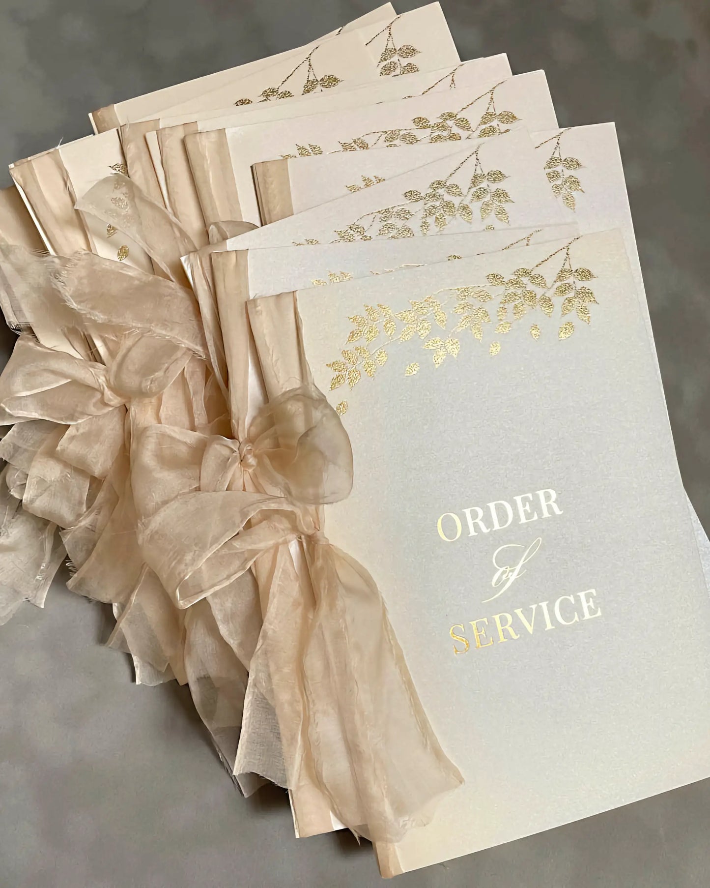 A stack of luxury wedding Order of Service booklets from the Foliage collection, featuring shimmering ivory paper with gold foil botanical designs and elegant peach organza ribbons — Polina Perri