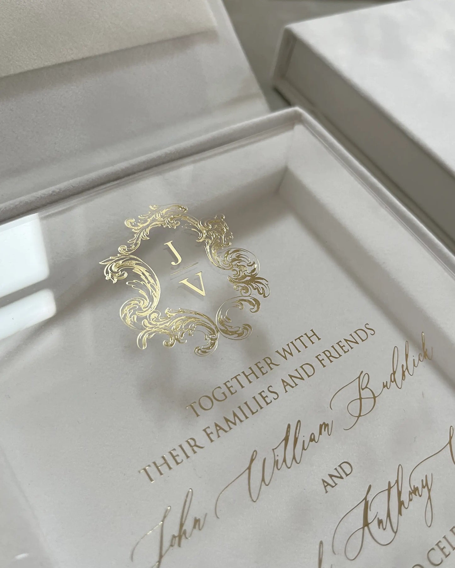 White Velvet Boxed Hampton Manor wedding invitation monogram by Polina Perri, macro shot of shiny gold foil stamped crest with initials JV on clear acrylic surface