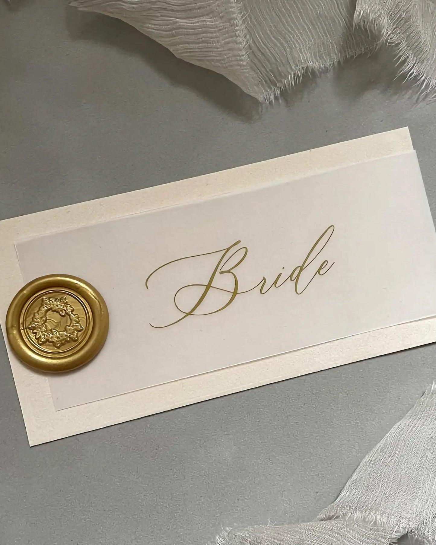 Elegant layered wedding place card featuring a translucent vellum overlay with gold foil script typography and a metallic gold wax seal on a textured background — Polina Perri