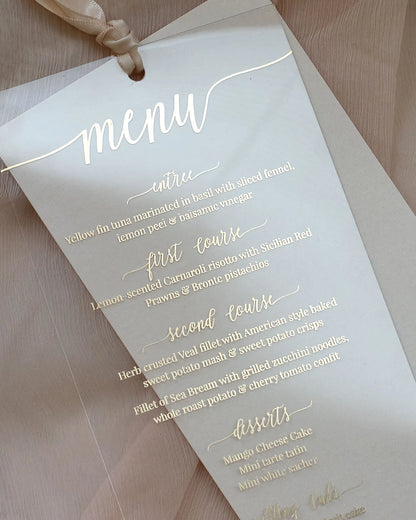 Close-up of a premium transparent acrylic wedding reception menu detailing courses in fine gold hot foil calligraphy over a neutral linen fabric — Polina Perri