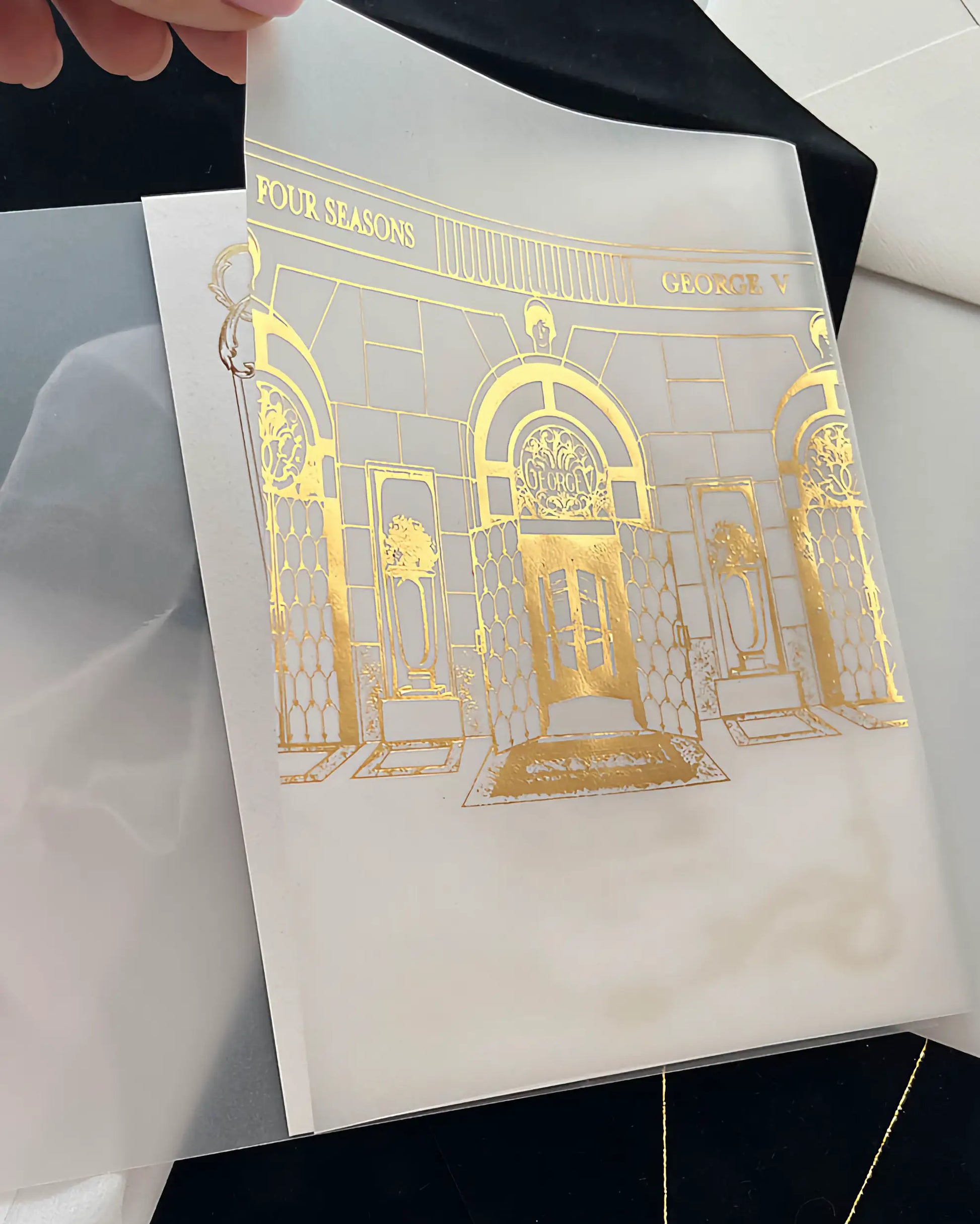Hand holding the translucent wedding invitation overlay showing the intricate metallic gold foil printing of the Four Seasons George V Paris gates — Polina Perri