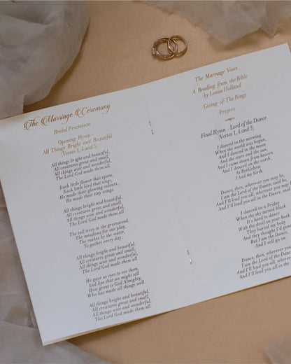 The inner layout of a wedding ceremony booklet showing printed hymns, order of events, and reflective gold foil headings next to a pair of wedding rings — Polina Perri