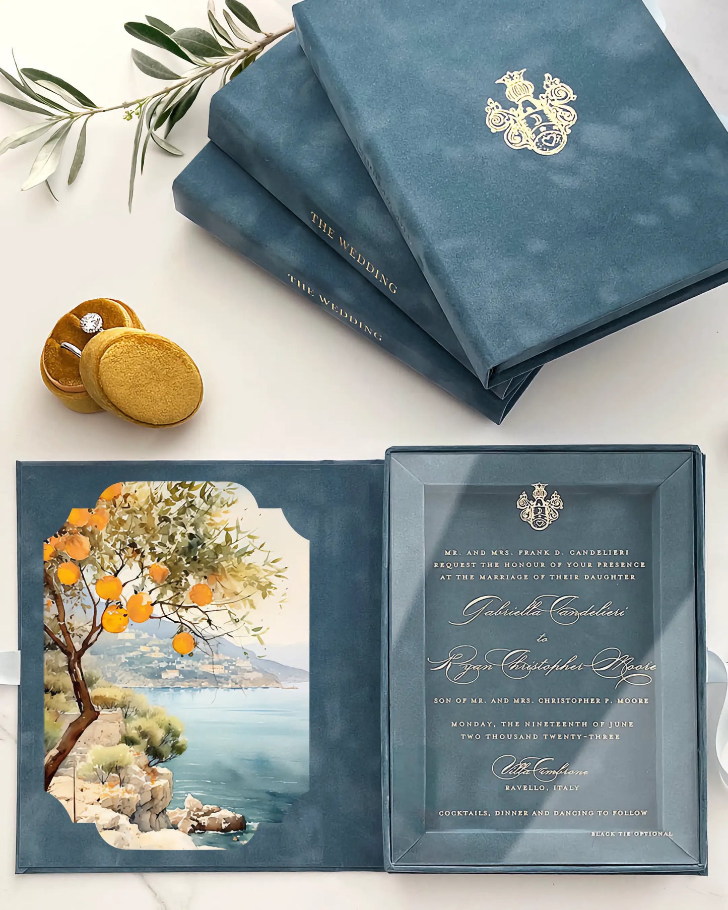 A luxury Italian destination wedding invitation suite featuring a dusty blue velvet box folio embossed with a gold monogram, opened to reveal a clear acrylic invitation card and a vibrant watercolor liner illustrating the Amalfi Coast with orange trees — Polina Perri