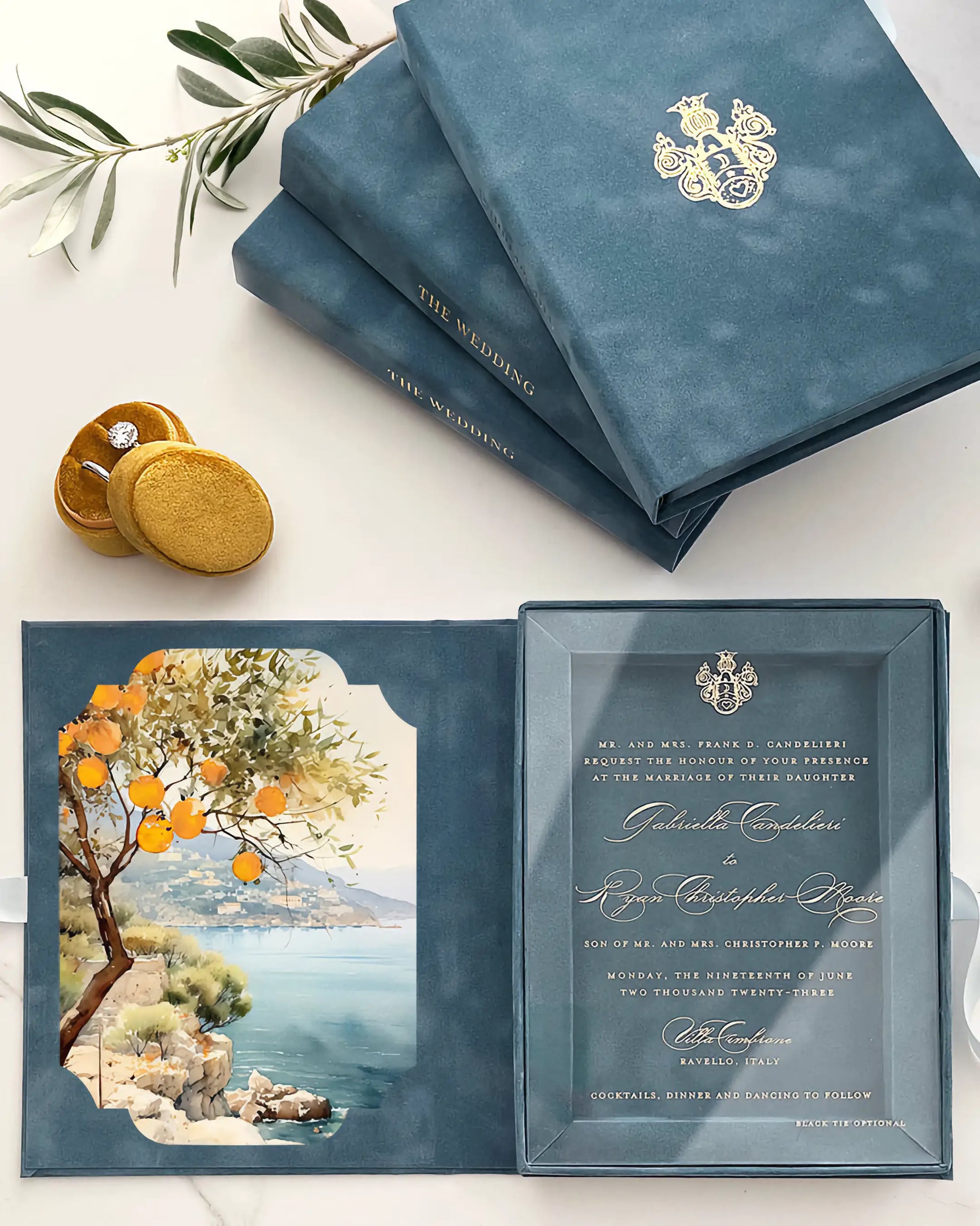 A luxury Italian destination wedding invitation suite featuring a dusty blue velvet box folio embossed with a gold monogram, opened to reveal a clear acrylic invitation card and a vibrant watercolor liner illustrating the Amalfi Coast with orange trees — Polina Perri