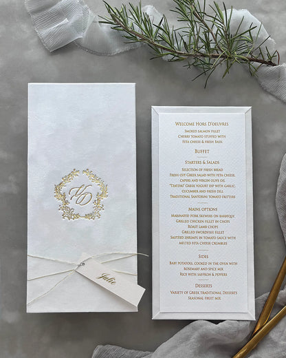 Minimalist white velvet wedding menu card ensemble with a gleaming gold hot stamped emblem cover, ivory cardstock insert, and fresh greenery accents — Polina Perri